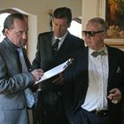 Drei Männer in Anzügen stehen in einem eleganten Raum. Einer schreibt auf einem Klemmbrett. Ein Gemälde ist an der Wand.