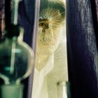 Ein Auge schaut durch ein zerschmettertes Fenster, umrahmt von violetten Vorhängen, mit unscharfen Glasobjekten im Vordergrund.