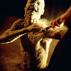 Verwitterte Statue taucht aus Sand auf, während eine Hand mit einem Werkzeug den Staub von ihr entfernt.