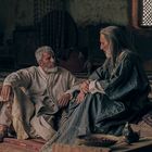 Zwei ältere Männer sitzen auf einem Teppich, tief ins Gespräch vertieft. Die Umgebung wirkt historisch und orientalisch.