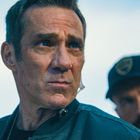 Ein Mann in einer blauen Jacke schaut ernst; im Hintergrund ein weiterer Mann mit Mütze und Bart.
