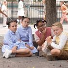 Auf einer Pflasterfläche sitzen vier Kinder nachdenklich zusammen, umgeben von spielenden Kindern im Hintergrund.