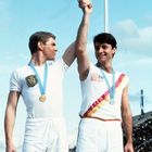 Zwei Sportler mit Medaillen: links ein Wappen auf dem Shirt, rechts US-Flaggen-Emblem. Beide heben Hände, Stadion im Hintergrund.