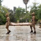 Zwei Personen in historischer Militärkleidung tragen eine Trage durch einen regnerischen Garten mit Bäumen und einem Gebäude.