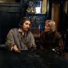 Zwei Männer in historischer Kleidung sitzen an einem Holztisch, darunter eine Ofenkachel mit blauen und gelben Mustern.