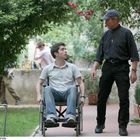 Ein Mann im Rollstuhl und ein Priester in schwarzer Kleidung unterhalten sich draußen in einem grünen Garten.