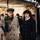 Ein junger Mann und eine Frau stehen auf einem Markt. Er trägt einen beigen Mantel und Mütze, sie einen dunklen Mantel.