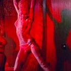 Keine erkennbaren Texte. Eine rote Beleuchtung zeigt eine tanzende Person und einen Trommler im Hintergrund.