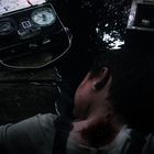 Ein Aufnahmegerät mit Tonband, blutender Verletzter in weißem Hemd liegt daneben, auf Holzboden, oben geöffnete Holzkiste.