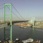 Im Vordergrund ist eine große Hängebrücke über einem Hafen zu sehen, umgeben von Kränen und Containern.
