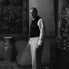 Ein Mann in einem Anzug steht in einem Raum mit antikem Mobiliar und kunstvollen Wandteppichen.