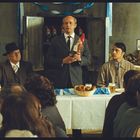 Links Schild: "Ich bin der 1. Gartenzwerg." Rechts Schild: "Ich bin der 100.000ste Gartenzwerg." Männer am Tisch, einer hält Zwerg.