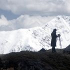 Eine Person mit Hut und Mantel steht mit einem Stativ auf einem Hügel vor schneebedeckten Bergen.