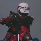 Auf einem weißen Helm steht "NAVA". Person auf Motorrad, trägt schwarzen gepunkteten Schutzanzug und hält den Lenker fest.