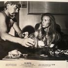 "THE SWAPPERS" unten. Ein Mann und eine Frau sitzen an einem Tisch mit Getränken, in einem Raum mit Jalousien. Schwarz-Weiß.