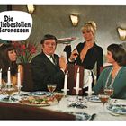 Text: "Die liebestollen Baronessen". Ein Mann und drei Frauen sitzen an einem gedeckten Tisch mit Kerzen und Wein.