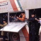 Der Text „Argos“ steht oben auf einem doppelstöckigen Bus. Menschen stehen mit Waffen in und außerhalb des Busses.