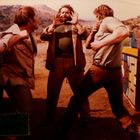 Drei Männer in Westernkleidung stehen auf einer staubigen Landschaft, ein Mann hebt beschwichtigend die Hände.