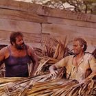 Zwei Männer in abgenutzter Kleidung stehen in einem Stapel aus Palmblättern vor einer hölzernen Wand und unterhalten sich.