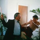 Zwei Männer in einem Kampf: Einer im Anzug mit Stock, der andere mit freiem Oberkörper beim Tritt. Pflanzen im Hintergrund.