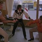 Texte: "DRUG" und "ANTIQUE". Drei Männer in einem Friseursalon, einer mit Gewehr, angespannte Atmosphäre.