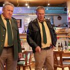 Zwei ältere Männer in grünen Uniformjacken und Krawatten stehen in einer Bar, dahinter ist ein Barkeeper in Weiß zu sehen.
