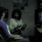 Zwei Personen sitzen in einem Raum. Eine hält eine Diskette, die andere ein Notizbuch. Ein Fernseher zeigt einen Nachrichtensprecher.