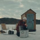 Eine Holzhütte mit blauem Türschild "OFFICE", zwei Personen fischen auf einem zugefrorenen See, Stühle und Ausrüstung daneben.