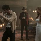 Drei Personen in eleganter Umgebung. Links macht jemand eine Handbewegung, rechts steht eine Frau in weißem Kleid, in der Mitte ein Mann mit Hut.