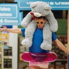 Text "Video Games & Games". Ein Mann balanciert mit ausgebreiteten Armen, einem riesigen Plüschbär auf den Schultern.