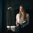 Eine Frau mit langen Haaren hält ein Notizbuch in einem abgedunkelten Raum mit Stehlampen. Sie trägt elegante Kleidung.