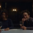 Zwei Personen sitzen an einem Tisch mit Weingläsern in einem gedimmten Raum, sie wirken entspannt und lächeln.