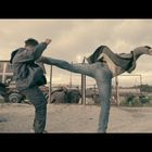 Ein Mann tritt gegen einen anderen, während zwei Personen am Boden liegen. Im Hintergrund ist eine verlassene Industrieanlage.