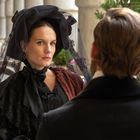 Eine Frau in schwarzer Kleidung sitzt unter Säulen, ihr gegenüber ein Mann in dunkler Jacke. Elegante, ernste Atmosphäre.