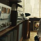 Ein Mann mit Kopfhörern kniet im Raum, vor ihm Audiosysteme. Im Hintergrund ein goldener Schallplattenrahmen.