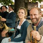 Mehrere Musiker in traditioneller Kleidung, entspannt sitzend, halten Blasinstrumente in einer ländlichen Umgebung.