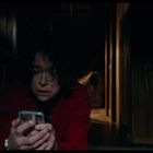 Eine Frau in rotem Mantel hält ein Smartphone fest, wirkt besorgt, im Hintergrund Bücher und ein dunkler Flur.