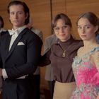 Eine Frau in Brille umarmt zwei junge Erwachsene elegant gekleidet, während Leute im Hintergrund klatschen.