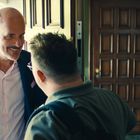 Ein Mann mit Glatze lächelt freundlich an einer geöffneten Tür. Ein anderer Mann steht mit dem Rücken zur Kamera.