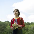 Ein Kind mit einem roten Pullover hält ein Notizbuch, steht vor einem Waldhintergrund unter einem blauen Himmel mit Wolken.