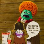 Ein Schild sagt: "You must be this tall to enter Big Guy Ride" vor einer grünen Figur mit orangefarbenem Afro.