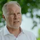 Keine Texte zu erkennen. Ein älterer Mann mit Bart in einem weißen Hemd, im Hintergrund Bäume.