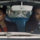 Zwei Personen sitzen in einem Auto. Die Frau hat kurzes Haar und trägt Schmuck, der Mann hat Dreadlocks und fährt.