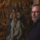 Ein Mann mit Brille und Bart sitzt vor einem Gemälde der Madonna mit Kind und umgebenen Figuren.