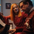Ein Mann und eine Frau in roter Kleidung am Klavier, unter einer Lampe. Notenblätter auf dem Klavier.