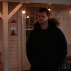 Ein Mann in Winterjacke steht auf einer Veranda mit Lichterketten, im Hintergrund ein Vogelhäuschen und eine Holzwand.