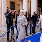 Zwei elegant gekleidete Frauen laufen auf einem blauen Teppich, umgeben von Fotografen. Sie tragen Sonnenbrillen und stilvolle Anzüge.