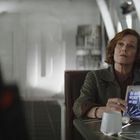 Eine Person sitzt in einem futuristischen Raum und hält ein holografisches Tablet mit komplexen Datenanzeigen.