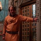 Ein Mann in einem orangefarbenen Anzug steht an einem verzierten, goldenen Aufzugsgitter in einem dekorativen Raum.