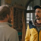Ein Mann in gelber Jacke und schwarzer Kappe steht in einem Raum mit Holzarbeiten und großen, bogenförmigen Fenstern. Ein anderer Mann mit grauem Pullover ist unscharf im Vordergrund zu sehen.